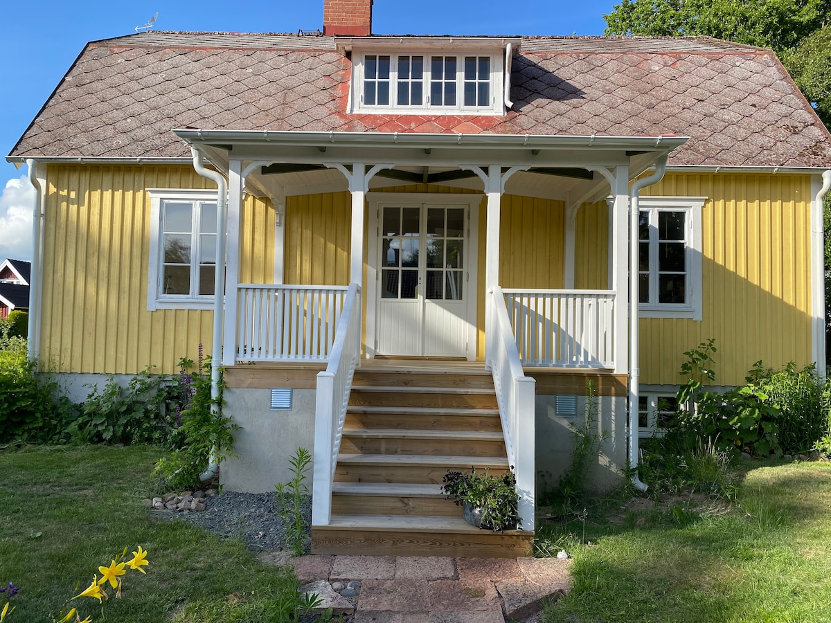 A charming yellow house features a welcoming front porch supported by white columns. Steps lead up to the entrance, framed by lush greenery and flowering plants. Large windows allow natural light to flood into the home, enhancing its inviting facade.