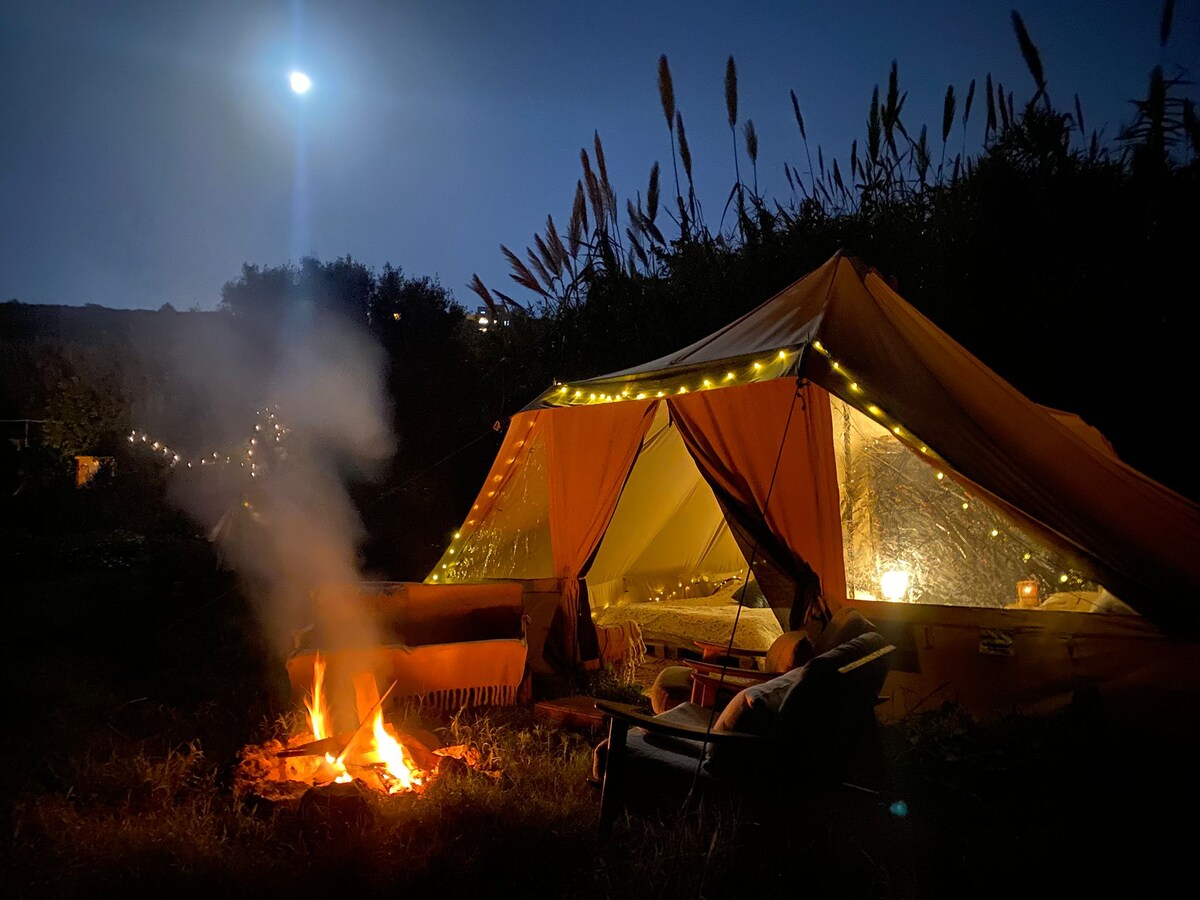 A cozy canvas tent is illuminated by warm lighting, set against a night sky with a full moon. A fire pit burns nearby, surrounded by seating. Lush greenery envelops the area, creating a serene camping atmosphere.