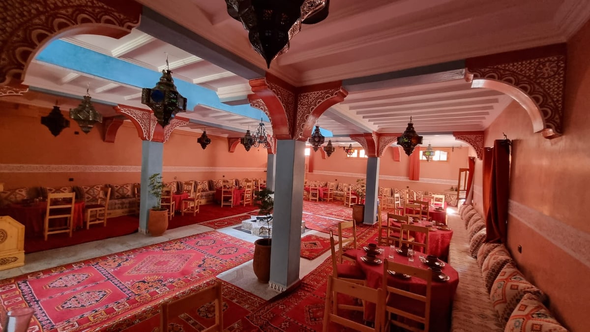 A spacious dining area is characterized by patterned carpets and cushioned seating along the walls. Intricately designed lanterns hang from the ceiling, while wooden tables are arranged throughout the space, offering a communal atmosphere complemented by warm hues on the walls.