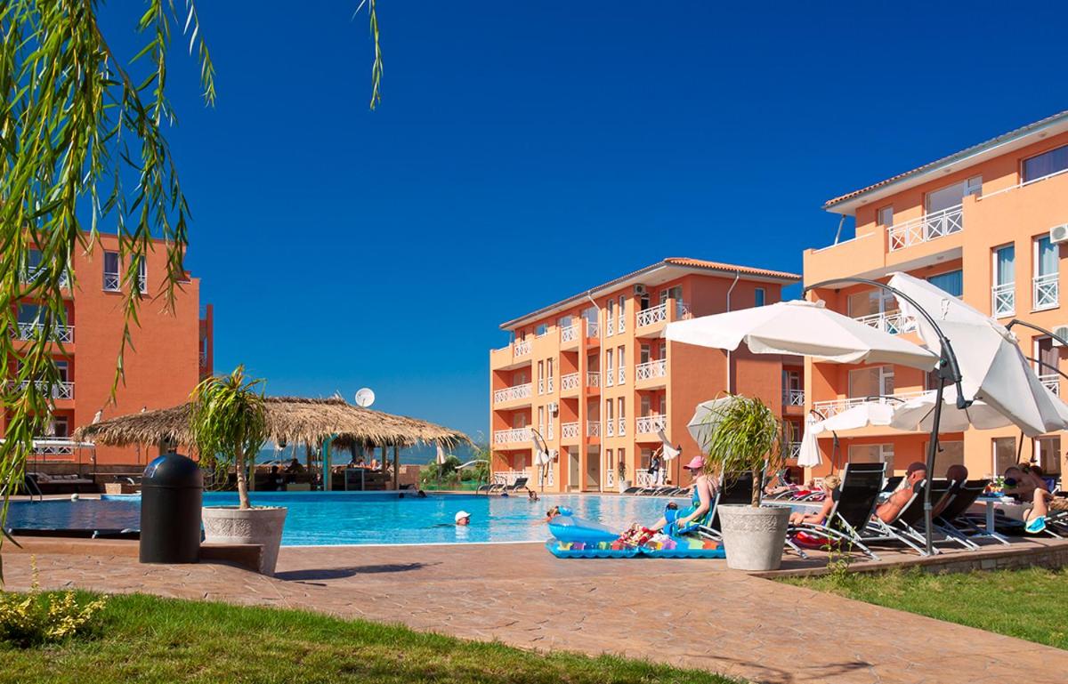 A serene pool area is surrounded by colorful buildings, featuring several loungers and umbrellas for shade. A thatched-roof cabana offers a shaded seating option. A clear blue sky enhances the vibrant atmosphere, while landscaped greenery adds a touch of nature.