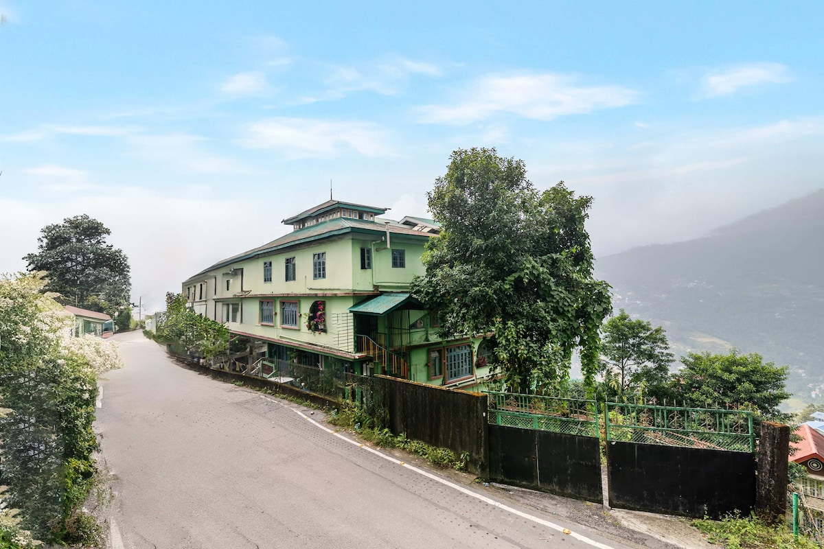 A charming homestay rises amid lush greenery, framed by the mountains of Gangtok. The building features multiple levels with balconies and expansive windows, inviting natural light. Surrounding trees and hedges complement the tranquil atmosphere, while the road curves gracefully, leading toward the inviting entrance.