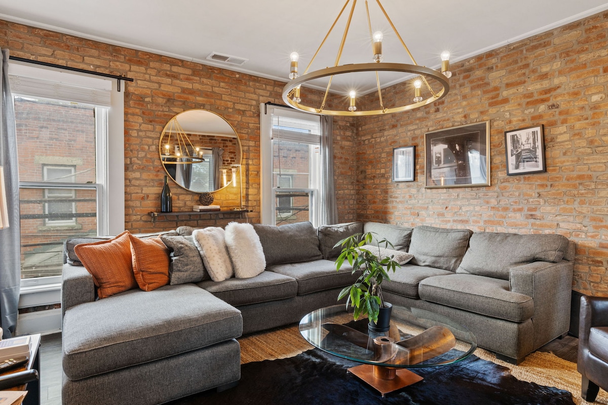 A cozy living room features a large sectional sofa adorned with decorative cushions. A round glass coffee table sits atop a dark area rug, complemented by a potted plant. Exposed brick walls and large windows create a warm ambiance, with a modern chandelier overhead.