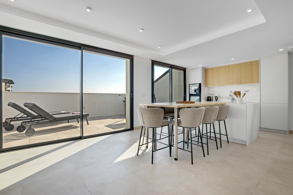 The kitchen and dining area feature a modern design with a large table and high-backed chairs. Large sliding glass doors create an open flow, allowing natural light to brighten the space and providing access to the outdoor terrace equipped with sun loungers.