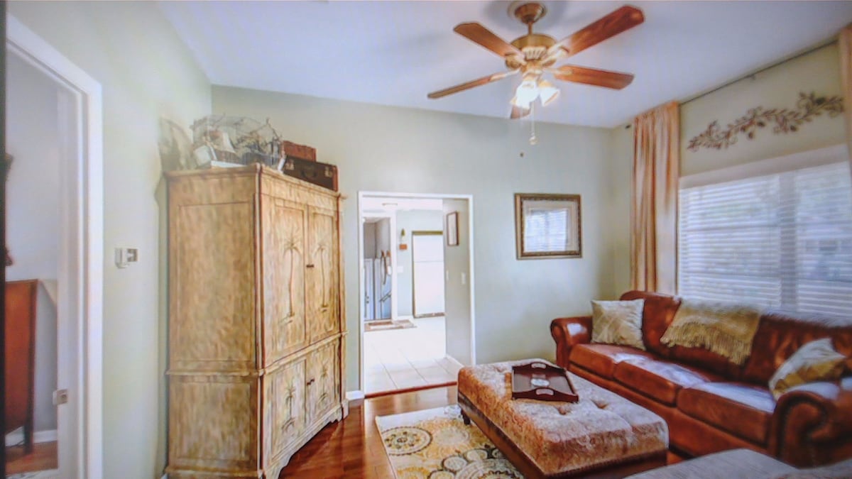 A living area is highlighted by a brown leather sofa and an ornate cabinet. A round ottoman rests on a patterned area rug, while a ceiling fan provides ventilation. Soft light filters through the window draped with light curtains, enhancing the space's warmth.