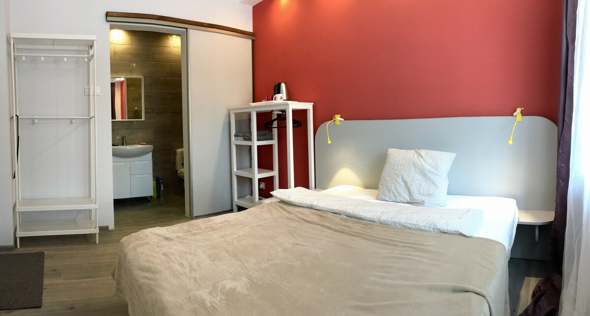 A spacious room is presented with a soft beige blanket draped over the bed. A red accent wall contrasts with the white furnishings, including a shelving unit and a hanging rack. A modern bathroom is visible through an open door, enhancing the room's functionality.