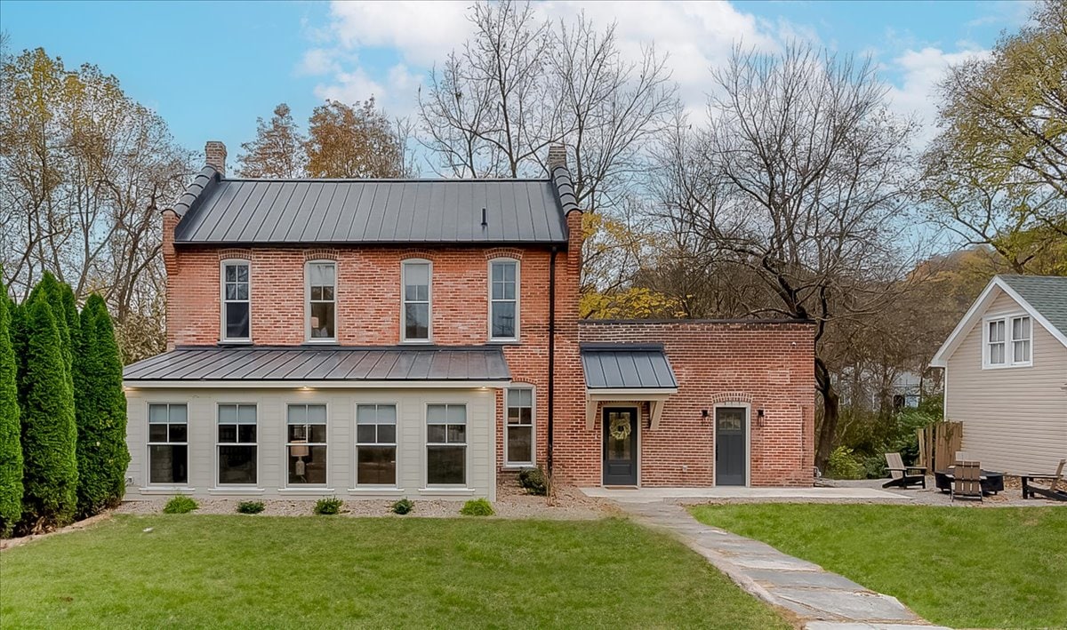 The historic carriage house features a two-story brick exterior with large windows offering abundant natural light. A pathway leads to the entry, framed by a well-maintained grassy area. Surrounding trees provide a natural backdrop, adding to the charm of the property.