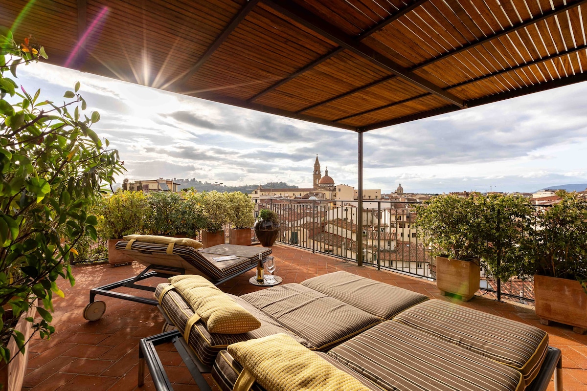 The spacious rooftop terrace features stylish designer deckchairs positioned for maximum comfort. Lush greenery surrounds the area, providing a tranquil setting with panoramic views of Florence's rooftops and iconic monuments. The sun casts a warm glow over the inviting space, creating a serene atmosphere.