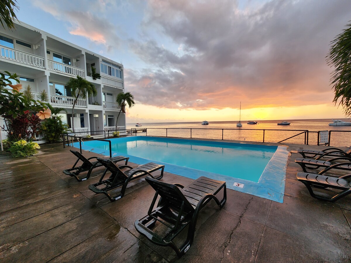 A serene pool area is situated beside the Caribbean Sea, featuring a rectangular pool framed by lounge chairs. The sky displays a gradient of colors at sunset, while boats are anchored in the distance. Two-story building with balconies overlooks the pool.