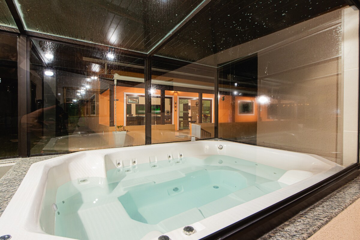 A spacious jacuzzi is enclosed in a glass-walled area, showcasing a serene ambiance. Soft lighting enhances the view of the bubbling water, with the inviting exterior visible in the background, framed by clean lines and muted colors.