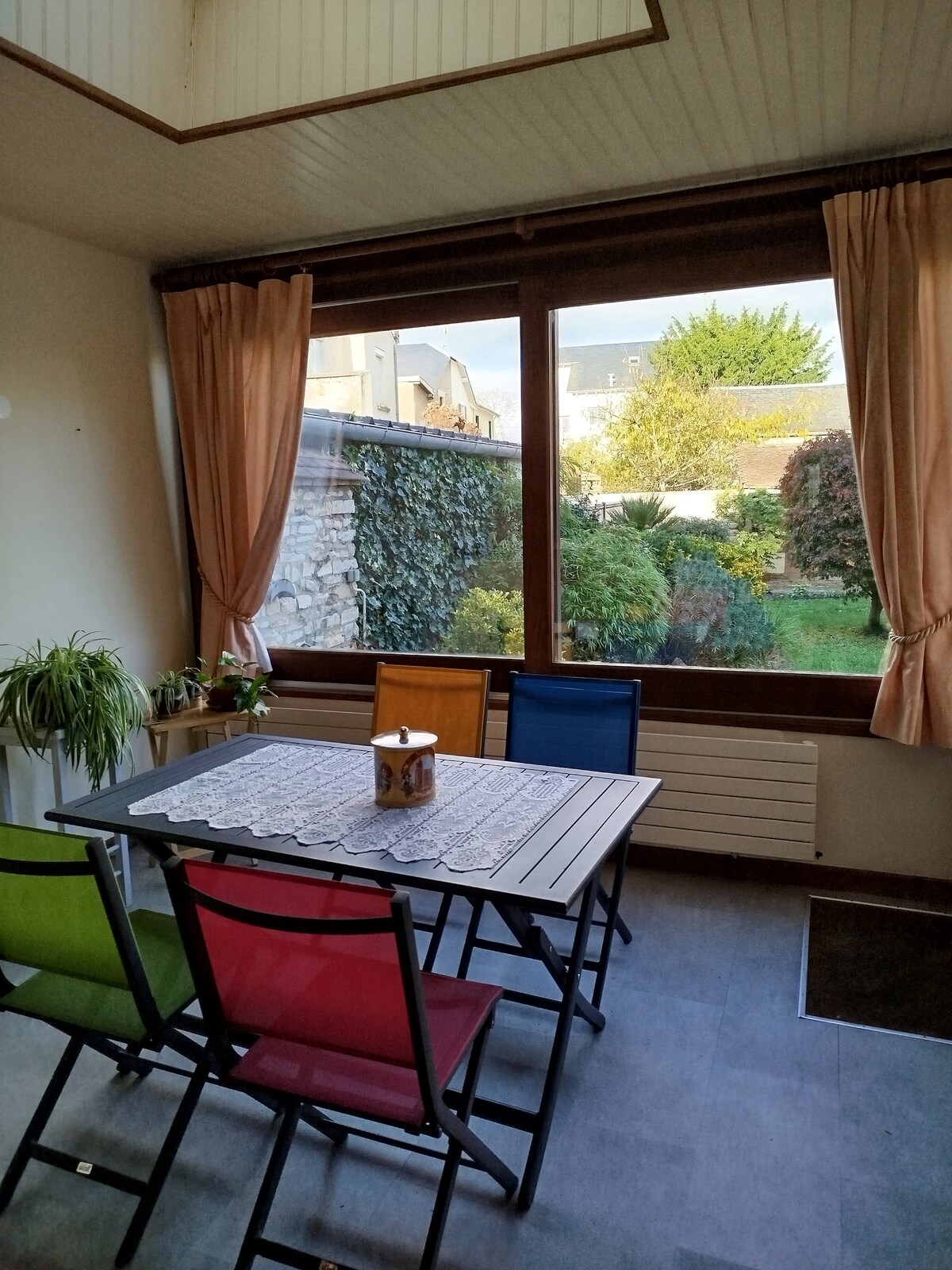 A dining area with a table and colorful chairs overlooks a garden through large windows. Natural light fills the space, highlighting the simplicity of the decor. Soft curtains frame the windows, offering a view of greenery and outdoor seating.