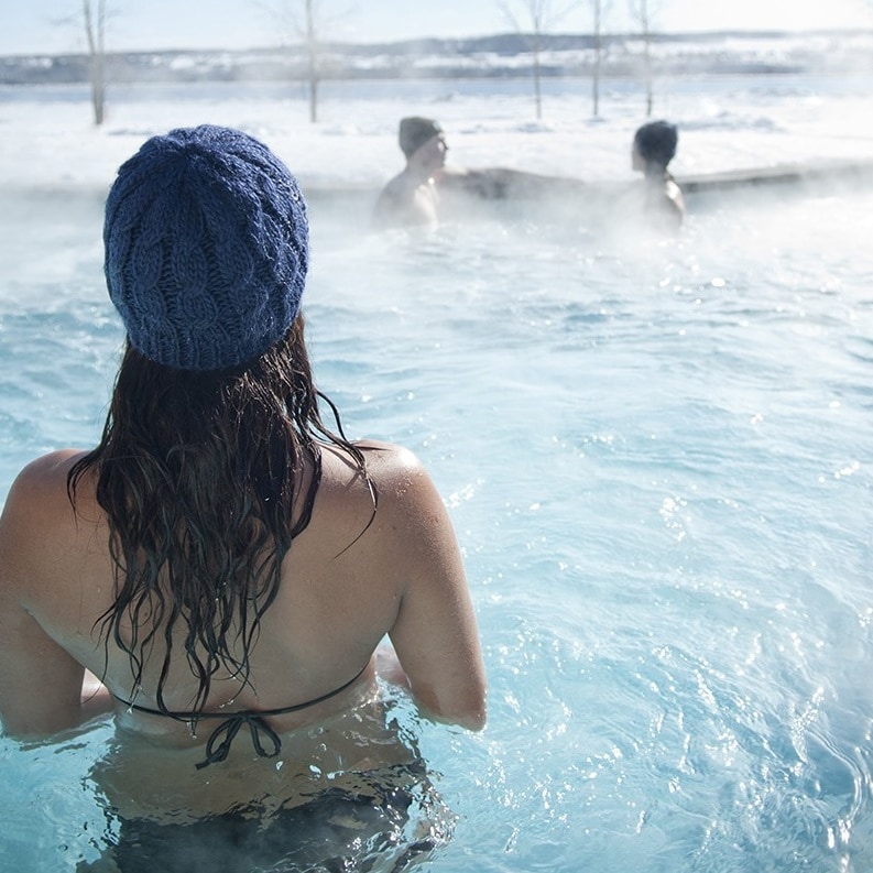 A serene outdoor spa scene is depicted with individuals relaxing in steaming, natural baths surrounded by a snowy landscape. Frost-covered trees are visible in the background, while soft steam creates a tranquil atmosphere.