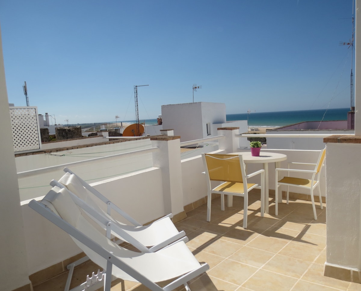 A private terrace is presented with views of the sea, featuring a small round table and two yellow chairs. Two reclining loungers are positioned nearby, creating a relaxing outdoor space. The clear blue sky complements the tranquil setting.