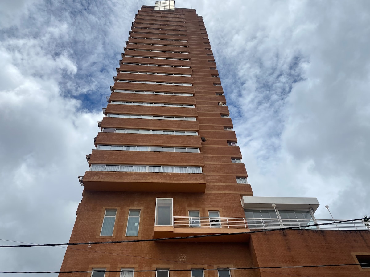 The exterior of a tall brick building is captured from below, featuring multiple balconies with sliding glass doors. A section of the building is highlighted with a large window at the top, under a partly cloudy sky.