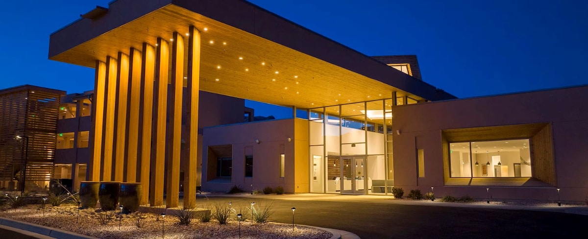The resort's entrance is illuminated at night, featuring modern architectural elements and a wooden canopy. Large glass doors provide a view into the lobby area, and neat landscaping with low-lying plants enhances the welcoming atmosphere.