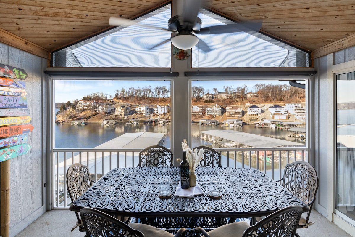 A spacious screened-in porch features a wrought-iron table with seating for six, overlooking serene lake views. Large windows provide natural light, and a ceiling fan adds comfort. Colorful signage decorates one corner, reflecting the lakeside charm of the surroundings.