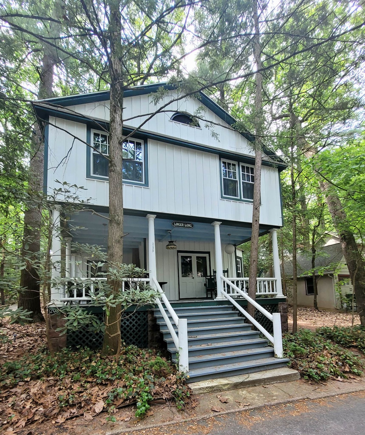 A charming two-story cottage is set among tall trees, featuring a welcoming front porch with white railings and steps. The exterior displays a light-colored siding with green accents, while surrounding foliage adds a natural touch to the peaceful ambiance.