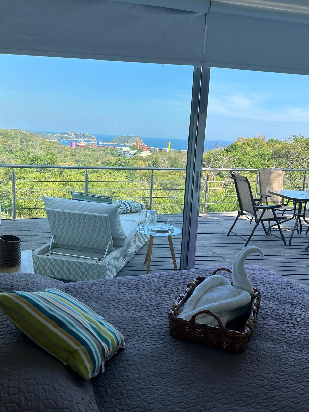 A spacious room features large glass doors that open to a deck with a sea view. An inviting seating area includes white lounge chairs and a table set for dining. Natural surroundings create a peaceful backdrop, enhancing the overall atmosphere.