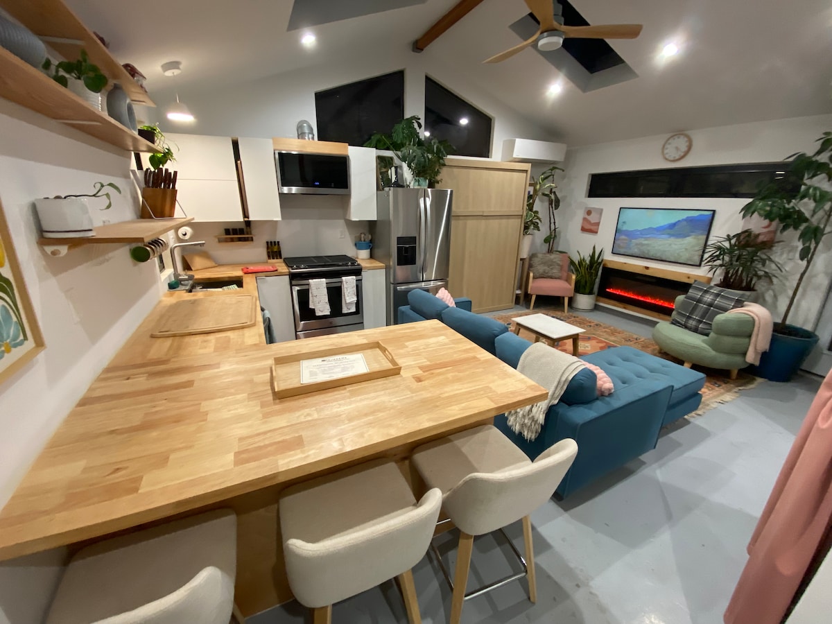 A modern kitchen and living area feature wooden counters and a spacious breakfast bar with cushioned stools. A sleek gas stove, stainless steel refrigerator, and open shelving are visible. Various plants add greenery, while a cozy seating arrangement is positioned near an electric fireplace and large screen.