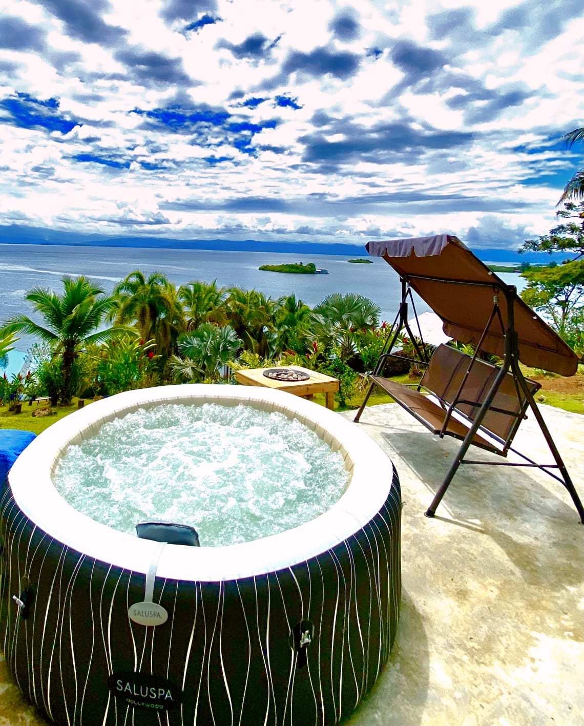 A jacuzzi filled with bubbling water is situated on a terrace overlooking a scenic body of water and lush greenery. A lounge chair is placed nearby, offering a space for relaxation while enjoying the expansive views. The sky is partly cloudy, enhancing the atmosphere.