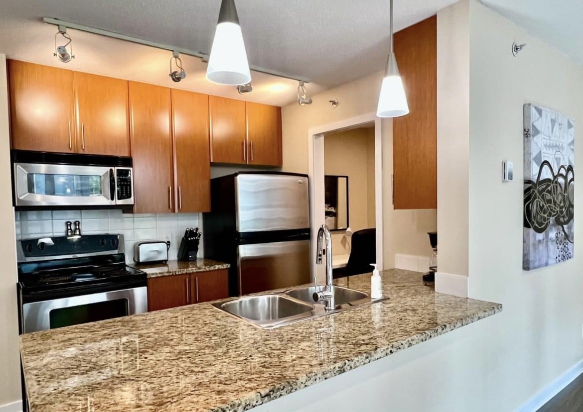 A well-equipped kitchen is presented, featuring modern appliances including a stainless steel refrigerator and stove. The granite countertops are complemented by wooden cabinetry. Two pendant lights hang above a double sink, creating a functional and inviting cooking space.