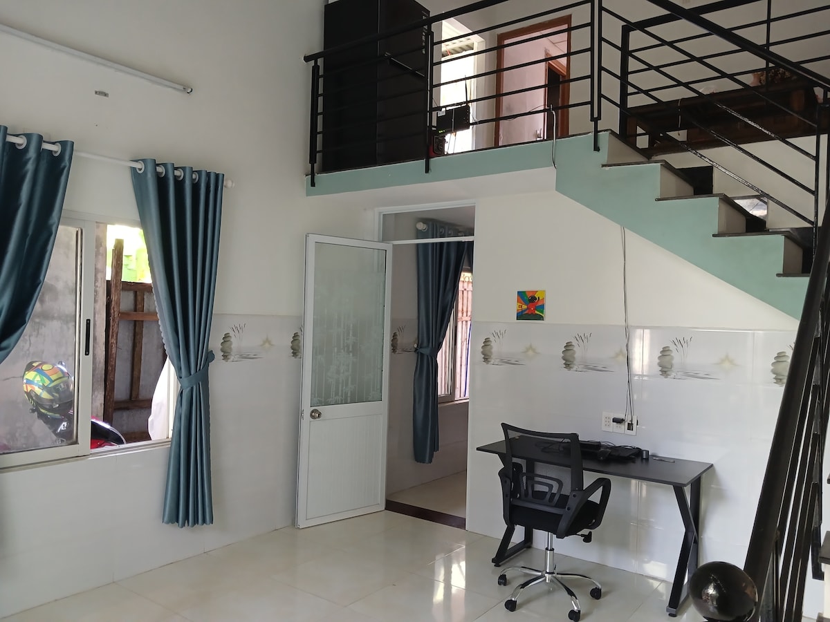 A well-lit interior features a modern workspace beneath a staircase. Large windows with light-filtering curtains allow natural light to enter. A colorful artwork is displayed on the wall, adding a touch of creativity to the space.