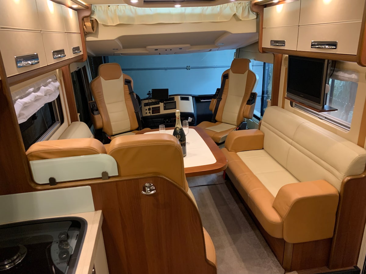 The interior of the motorhome features a comfortable seating area with light-colored leather chairs and a spacious sofa. A table is positioned at the center, with cabinetry and a small kitchenette visible in the background, providing a cozy and functional space for relaxation.