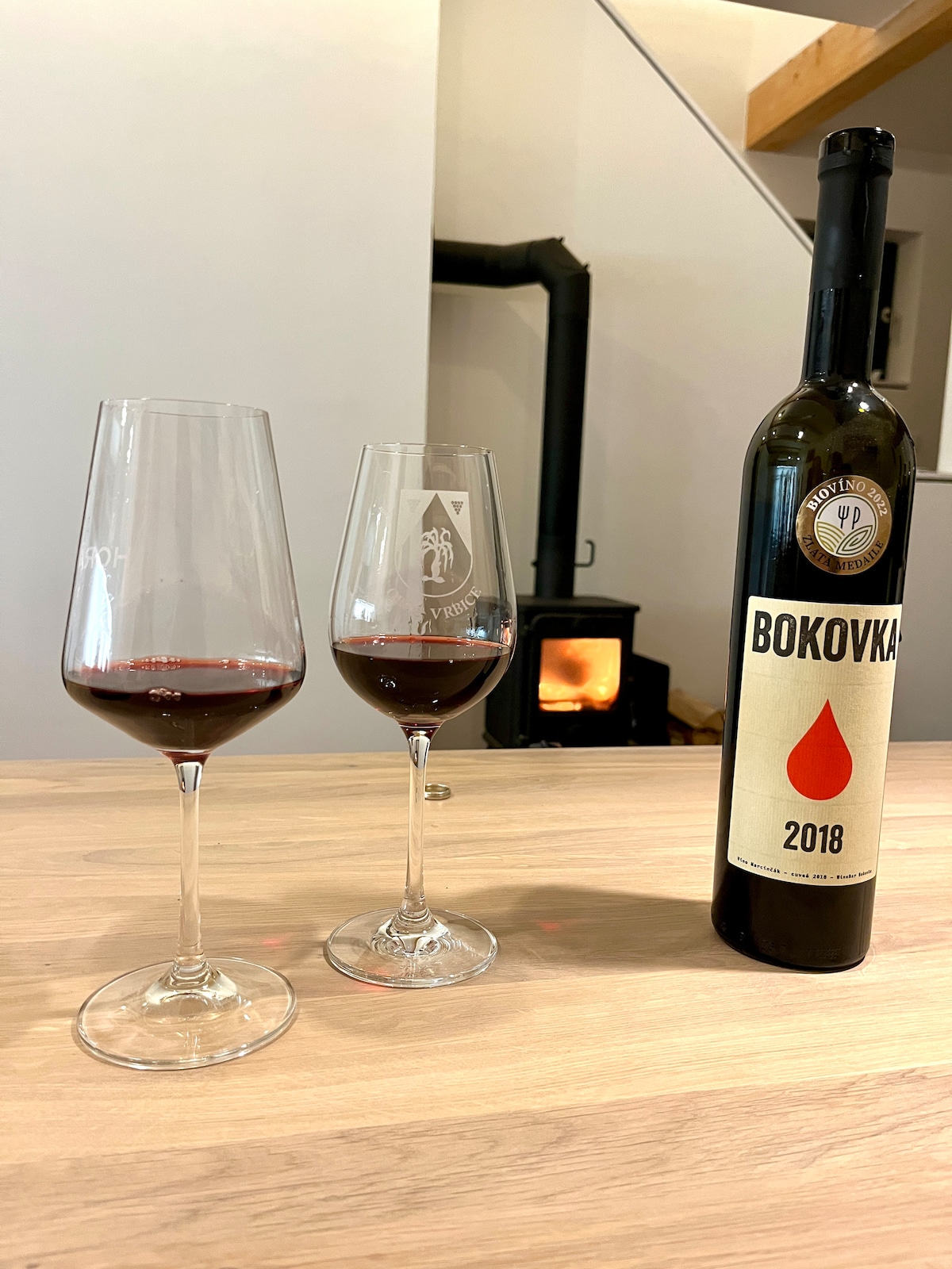 Two elegantly shaped wine glasses filled with deep red wine are placed on a light wooden surface. A bottle labeled 'BOKOVKA 2018' stands beside them. A warm glow from a nearby wood-burning stove creates an intimate ambiance in the setting.