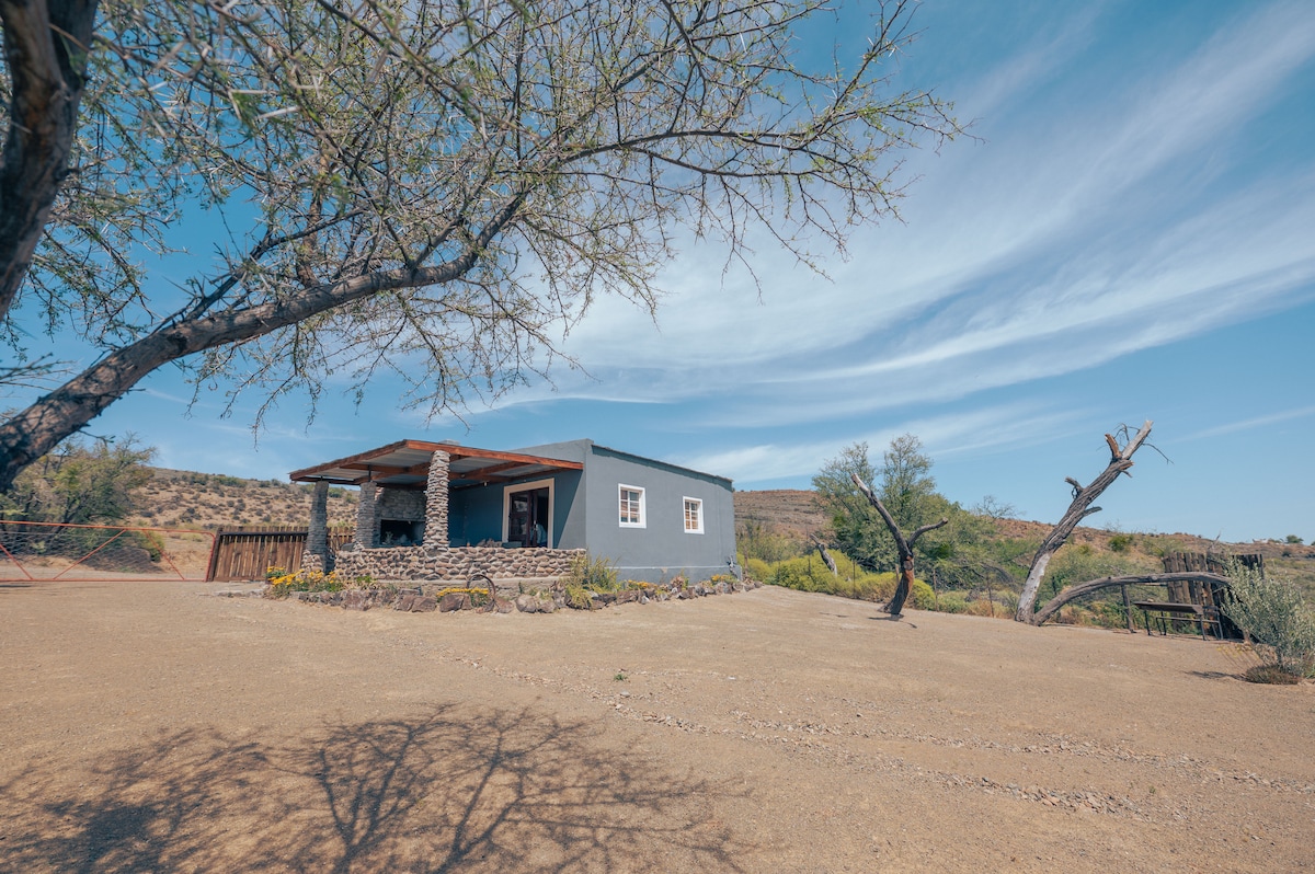 A cottage is portrayed against a clear blue sky, featuring a stone patio with a wooden awning. Surrounding the home are sparse trees and resilient shrubs, highlighting the natural landscape of the Grootwaterval Farm.