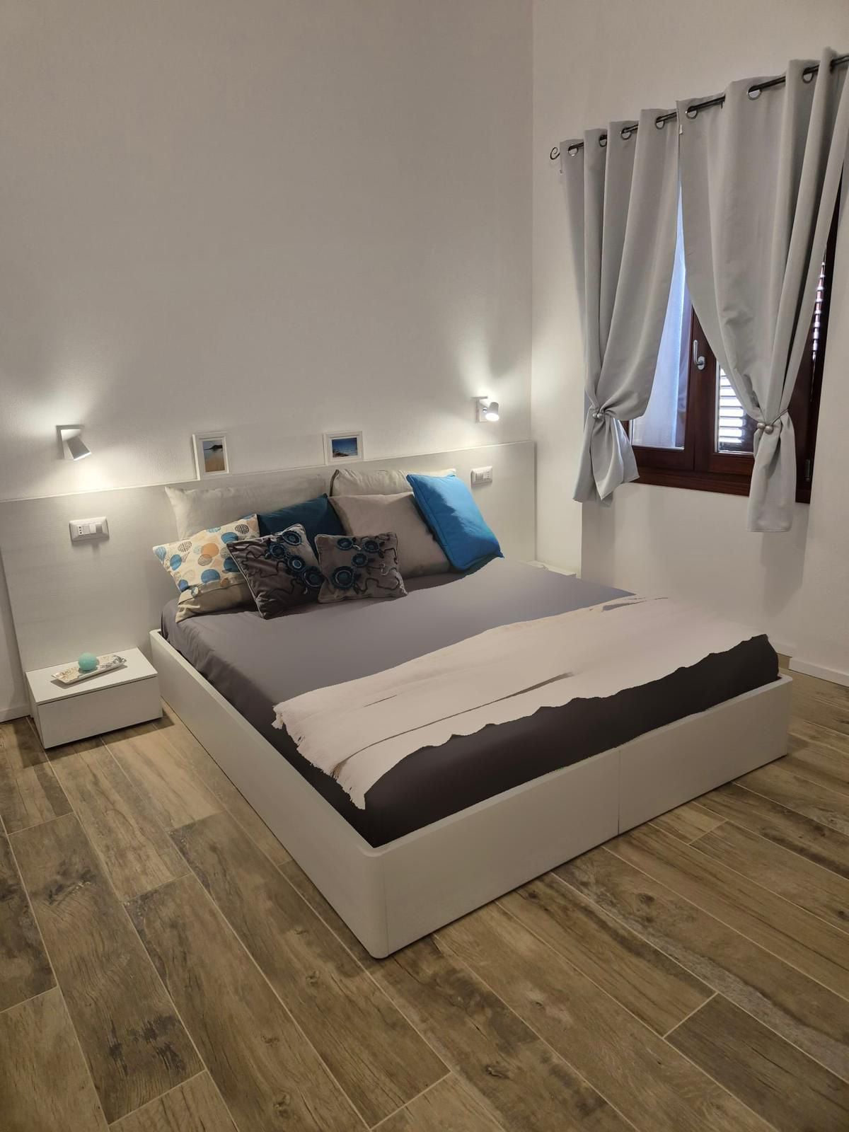 A comfortable bedroom featuring a spacious bed with neutral bedding and decorative pillows in various shades of blue. A bedside table holds a small lamp, and natural light filters in through partially draped windows, complemented by softly colored curtains.