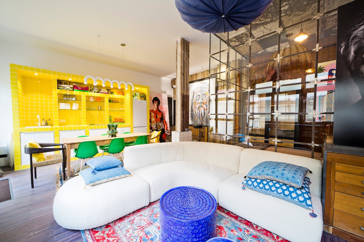 A spacious living area features a curved white sofa with decorative pillows, complemented by a vibrant blue table. Bright yellow cabinetry and a glass partition enhance the open layout, with a dining area visible in the background, showcasing green chairs and a small plant.