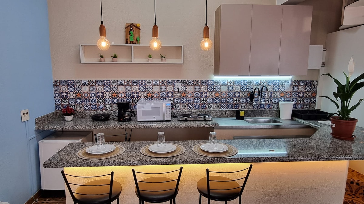 A modern kitchen area features a spacious countertop lined with three stools. Soft lighting highlights the decorative tile backsplash, complemented by essential kitchen appliances, including a microwave and coffee maker, arranged for convenient use.