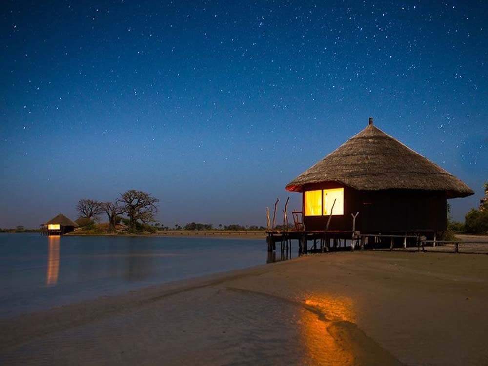 A thatched-roof hut sits on stilts by the water's edge, illuminated from within. The calm surface of the lagoon reflects the bright starry sky, creating a serene atmosphere. Another similar structure is visible in the distance, surrounded by a quiet natural landscape.