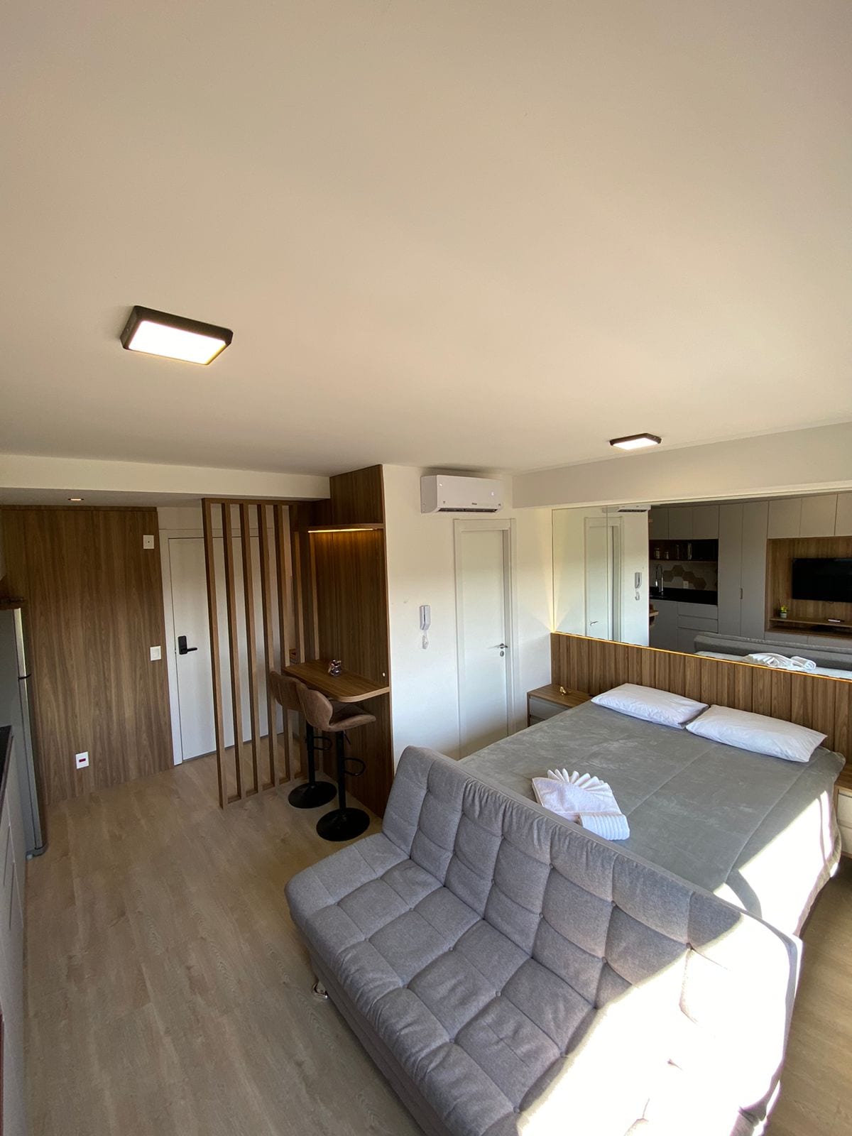 A modern studio layout features a comfortable sleeping area with a spacious bed and a cozy sofa. Light-colored wooden accents are present throughout. A small dining area is visible, and the kitchen area is equipped with essential appliances. Natural light fills the space from the window.