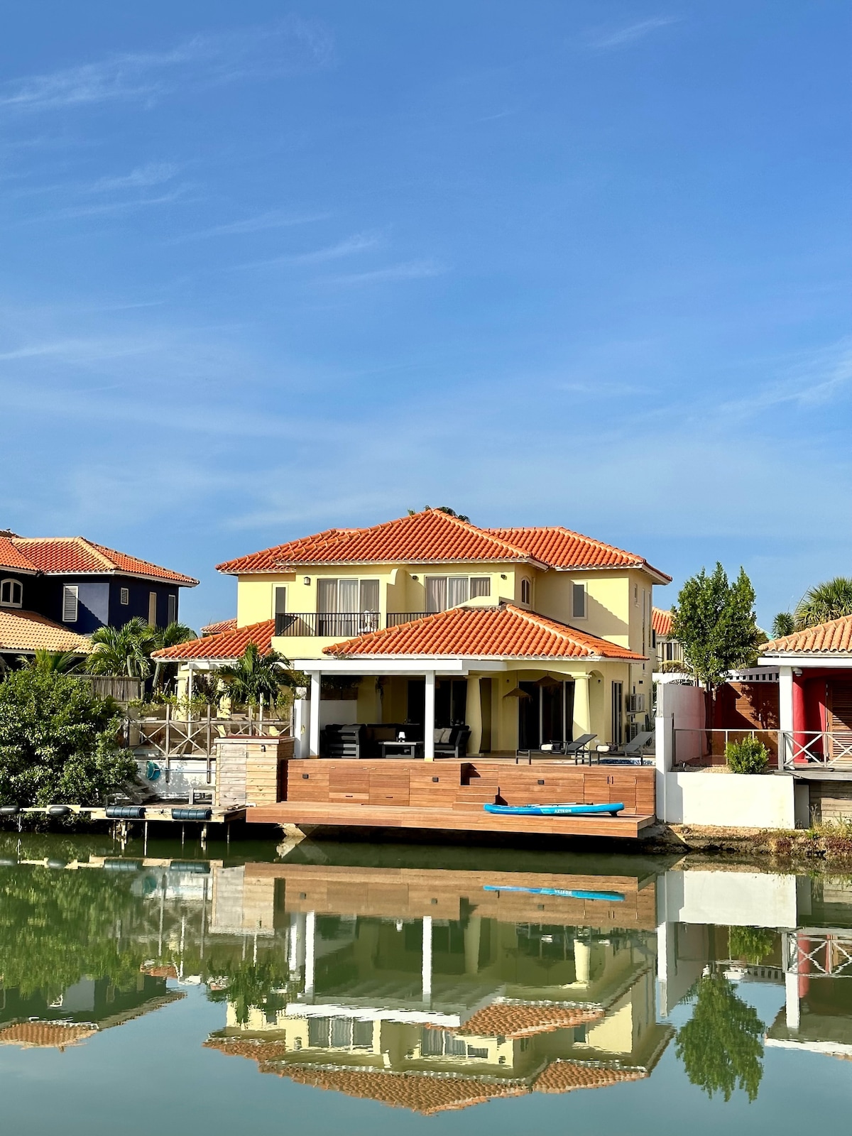 A charming villa is seen across the shimmering water, featuring a distinctive red-tiled roof and light yellow exterior. The structure has expansive glass doors opening to a deck, with surrounding tropical greenery enhancing the serene waterfront setting.