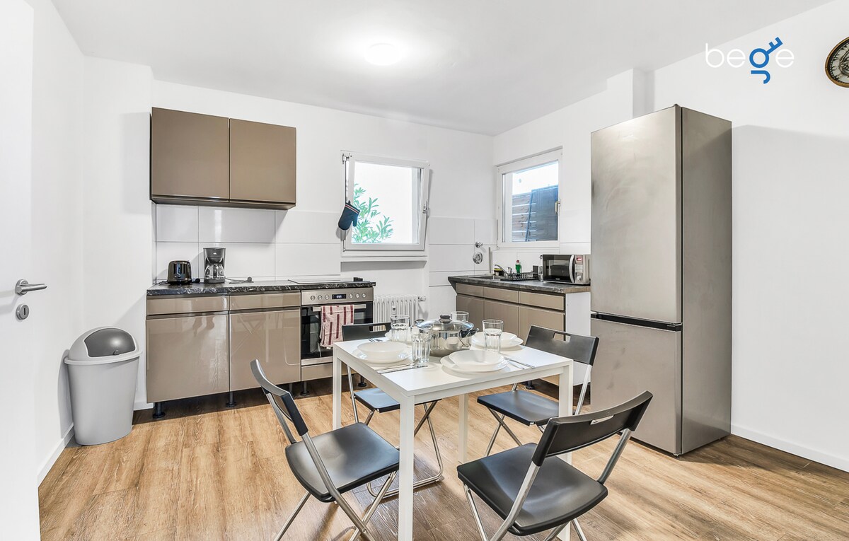 A modern kitchen is equipped with sleek cabinetry and stainless steel appliances. A dining table is set for four, complemented by simple chairs. Natural light enters through the windows, highlighting the functional layout and clean design of the space.