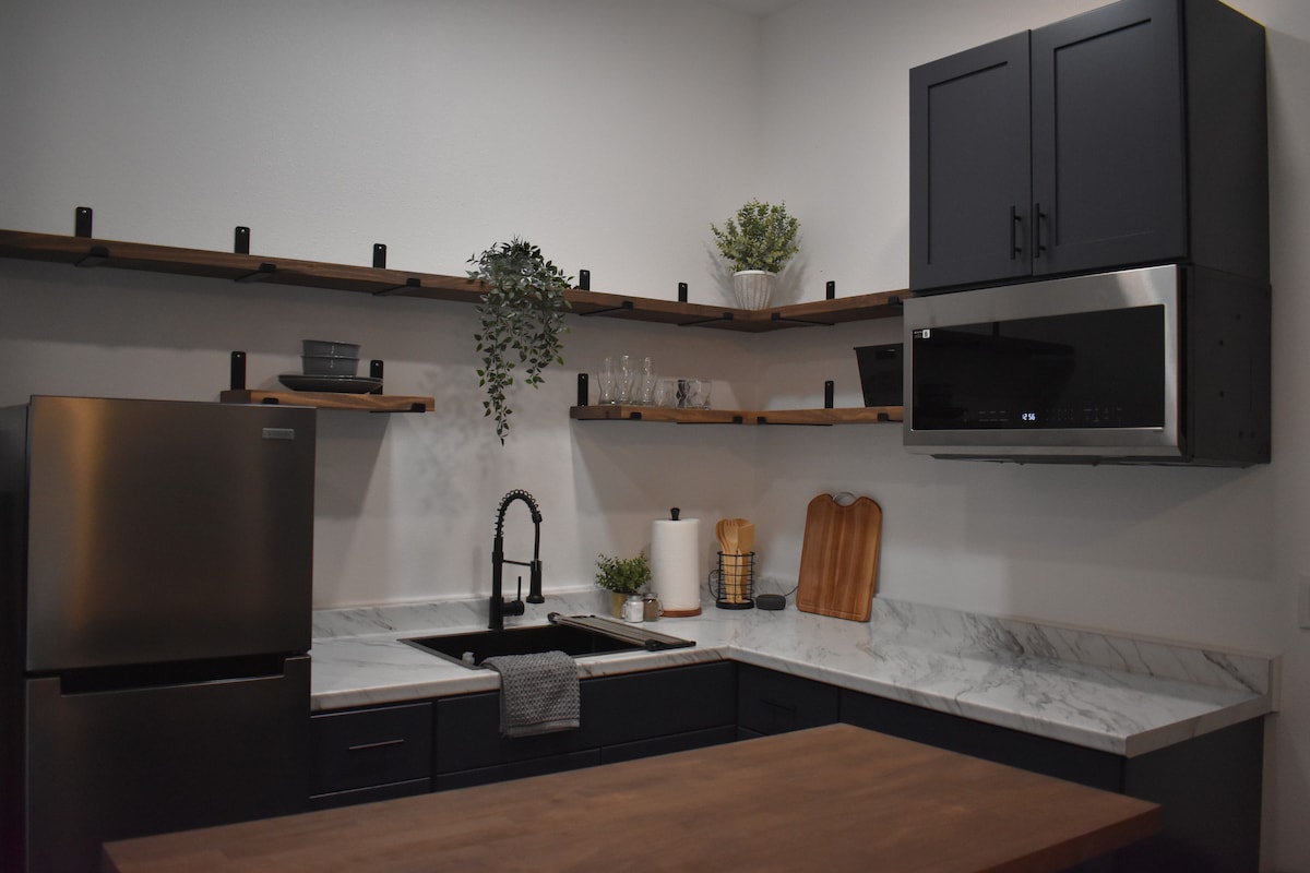 A modern kitchen features dark cabinetry complemented by open wooden shelves. A stainless steel refrigerator stands beside a sleek sink area, which is equipped with a faucet. Marble countertops provide workspace, while various kitchen utensils and decor items are neatly arranged on the shelves.