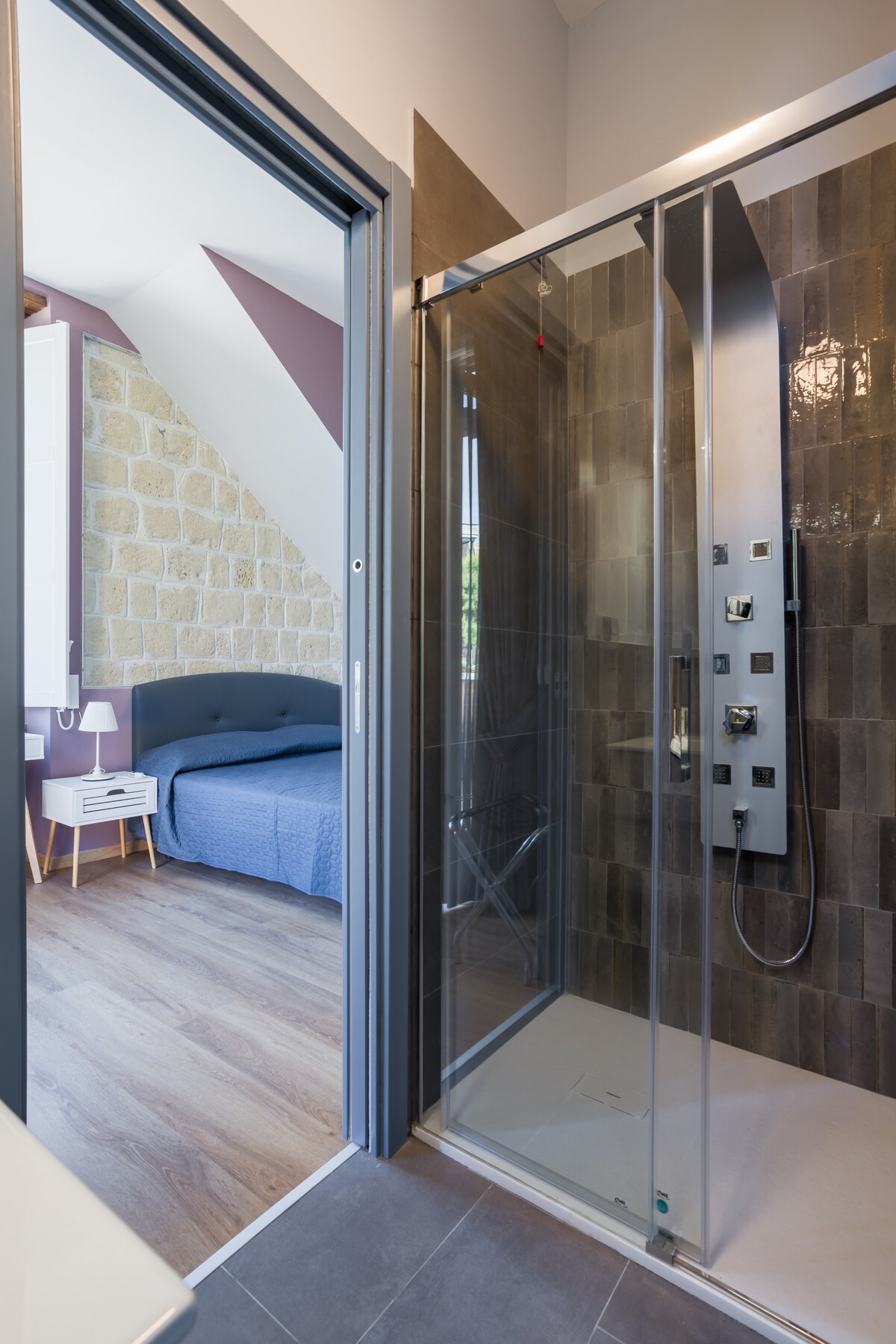 A modern bathroom features a glass-enclosed shower with sleek fixtures, displayed alongside a textured stone wall. The adjacent bedroom is visible, showcasing a spacious bed with a blue coverlet, complemented by a nightstand with a lamp.