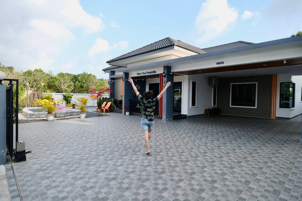 An outdoor area features a spacious paving stone surface, flanked by colorful plants. The entrance to the house is visible, showcasing a modern architectural style. A few lounge chairs can be seen in the corner, providing an inviting space for relaxation.