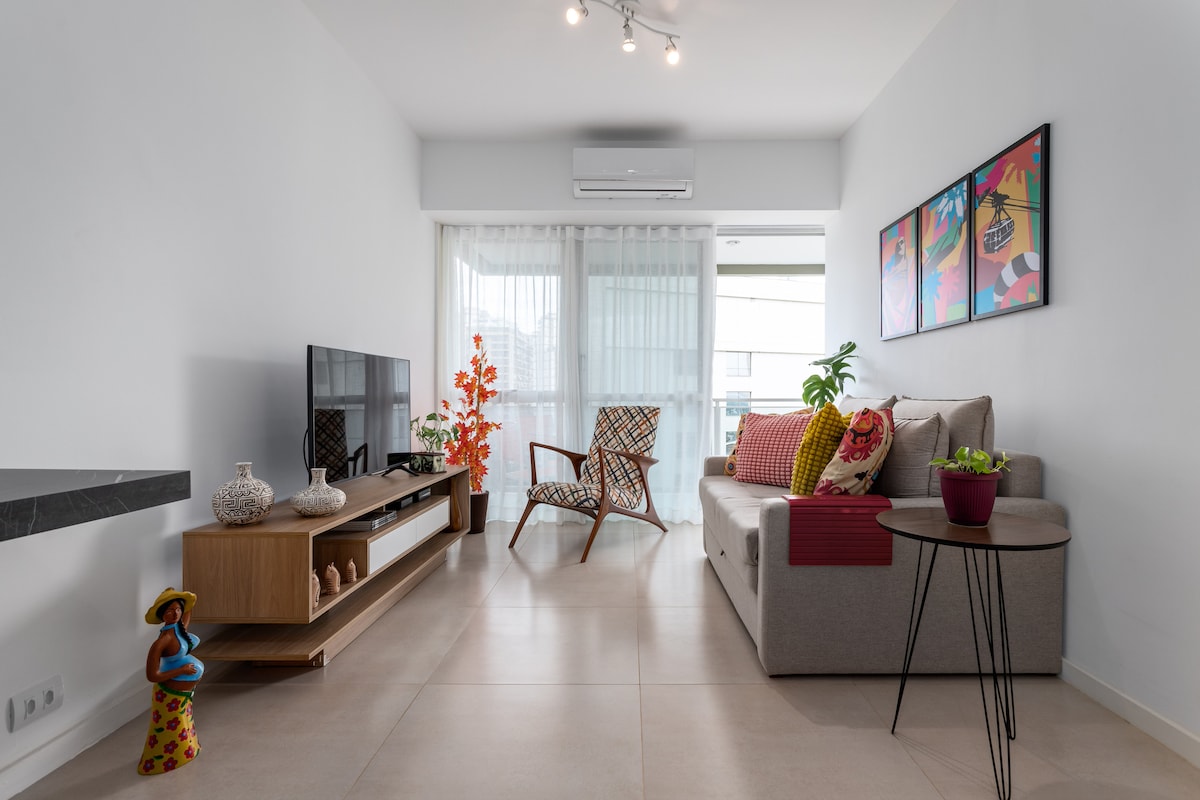 A modern living area features a comfortable sofa bed alongside a stylish chair. A flat-screen TV is mounted on a wooden unit, while decorative plants and artwork add a touch of color. Natural light streams in through large windows with sheer curtains.