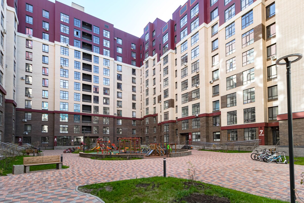 The courtyard of an apartment complex showcases a spacious layout with colorful playground equipment and benches. Surrounding greenery and paved walkways enhance the outdoor area, which is framed by the striking architecture of the surrounding buildings.