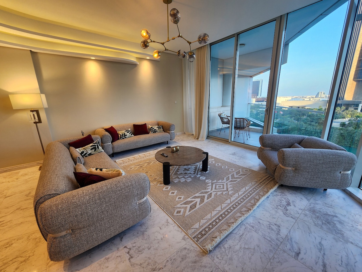 A modern living area features a spacious gray sofa adorned with patterned throw cushions. A circular coffee table sits at the center, complemented by a cozy armchair. Floor-to-ceiling windows provide views of the outside, allowing ample natural light to fill the space.