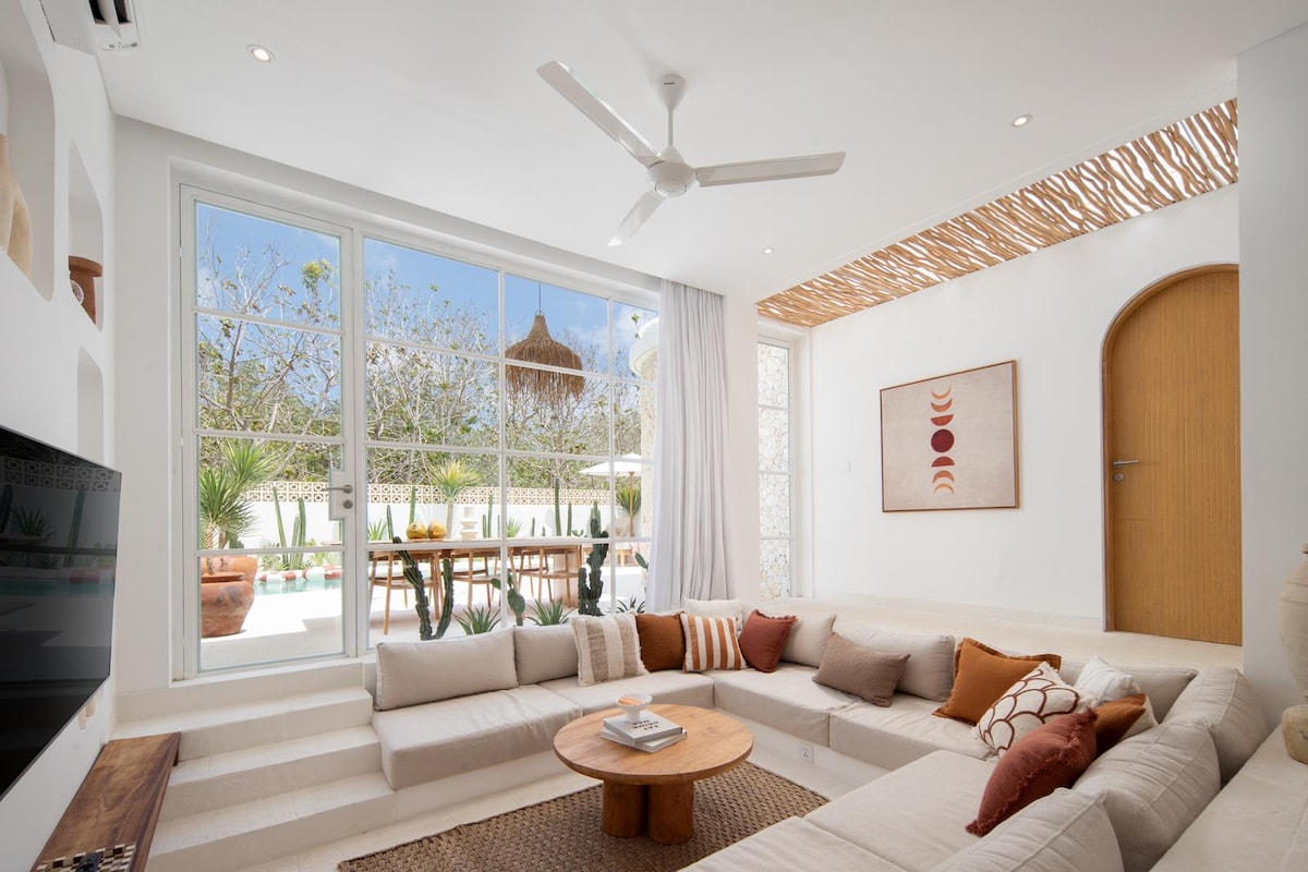 A sunlit living area features a large sunken lounge arranged with cream-colored cushions. A round wooden table is centered in front, surrounded by natural light filtering through expansive windows. Decorative elements include a woven ceiling and artwork on the wall, contributing to a relaxed atmosphere.