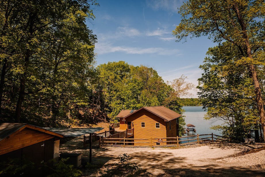 A cozy wooden house is nestled among lush trees, with views of the calm lake nearby. A gravel pathway leads to the residence, surrounded by open space and nature. The sun shines brightly, casting a warm light over the inviting outdoor setting.