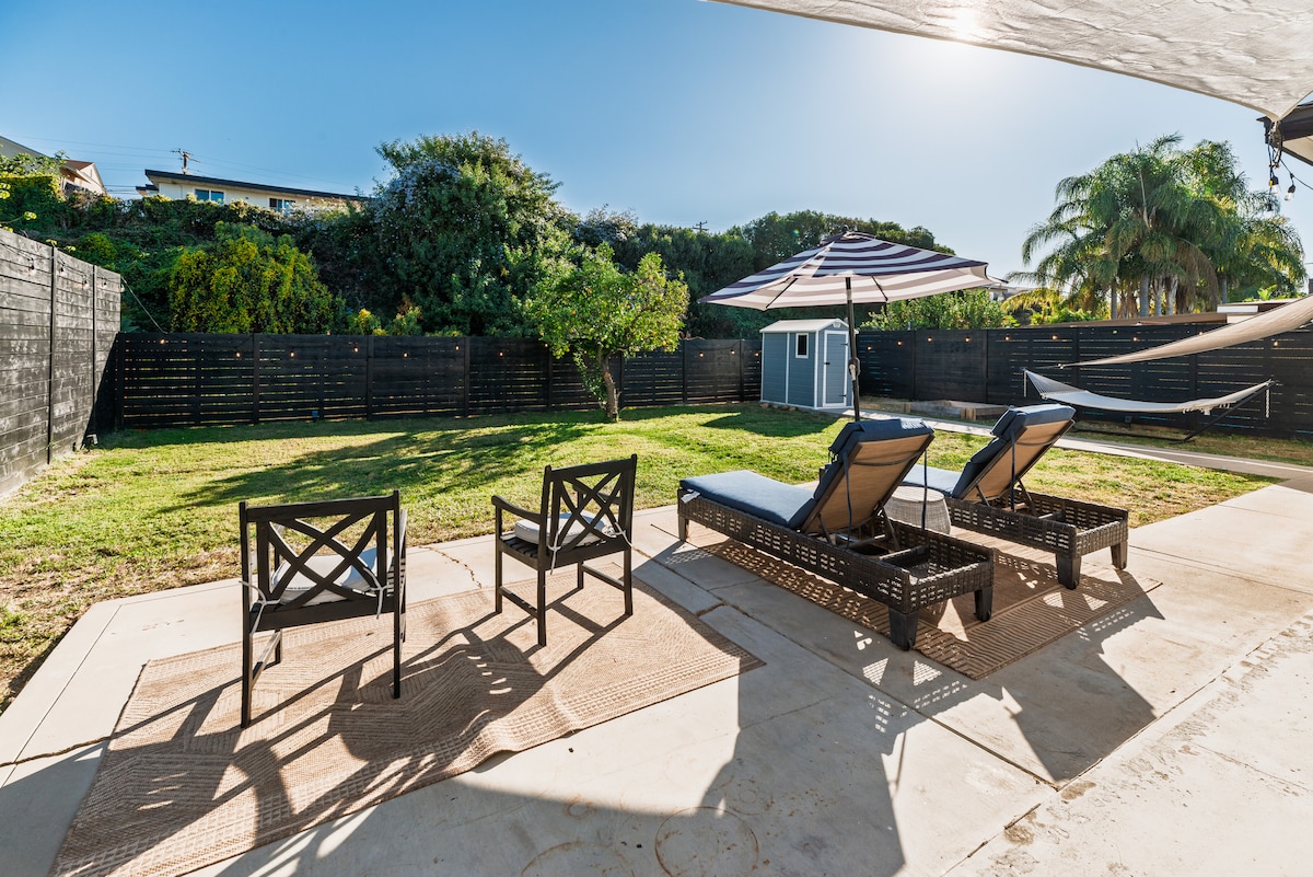 An expansive backyard area is visible, featuring two lounge chairs positioned on tan mats. A large umbrella provides shade over the chairs, and a hammock is seen in the background. Green grass covers the ground, bordered by a tall black fence and foliage.