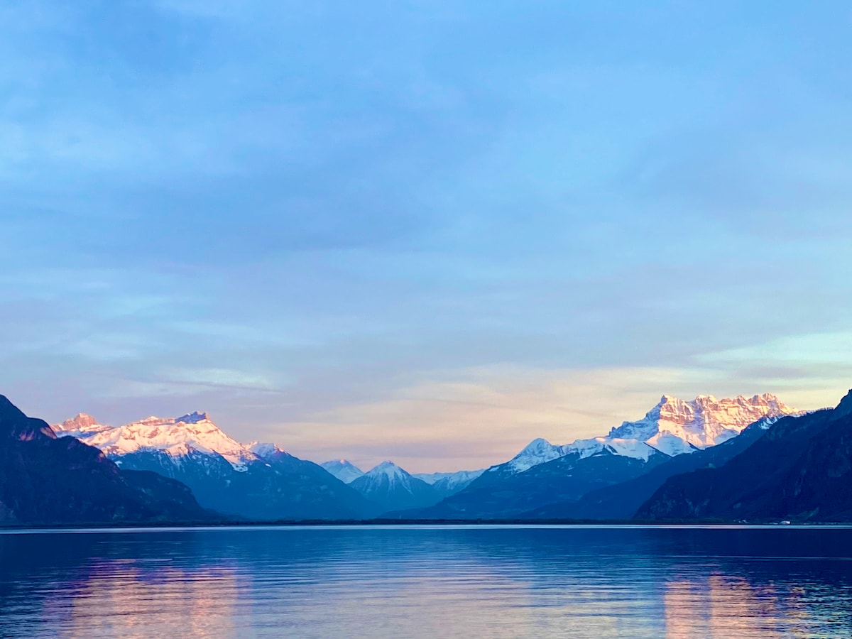A serene view of a lake is presented, with distant snow-capped mountains reflecting softly in the water. The sky transitions to shades of blue and pink, adding a gentle hue to the tranquil scene. The calm surface of the lake enhances the distant landscape.
