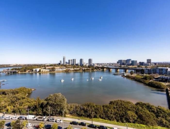 An expansive view of a serene waterfront is showcased, with gentle waves reflecting a clear blue sky. The foreground displays lush greenery along the shore, while modern buildings rise in the distance, framing the peaceful scene.