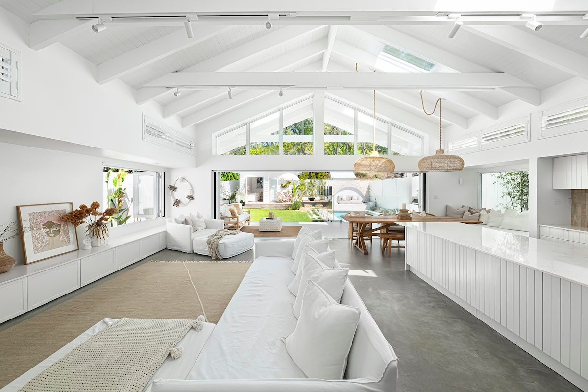 An open-concept living space features a high-pitched ceiling and large windows that invite natural light. A spacious white sofa with soft cushions is placed alongside a neutral rug. A wooden dining table is visible, complemented by pendant lighting, enhancing the modern aesthetic.