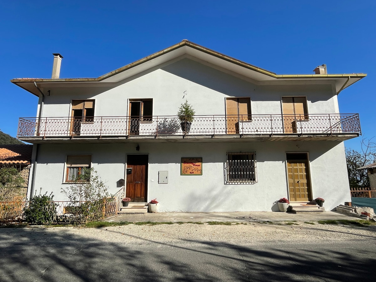 A two-story building is depicted, featuring a light-colored exterior and a sloped roof. A wide balcony extends across the front, with wooden shutters on the windows. The entrance door is framed by a small garden area, enhancing the tranquil setting.