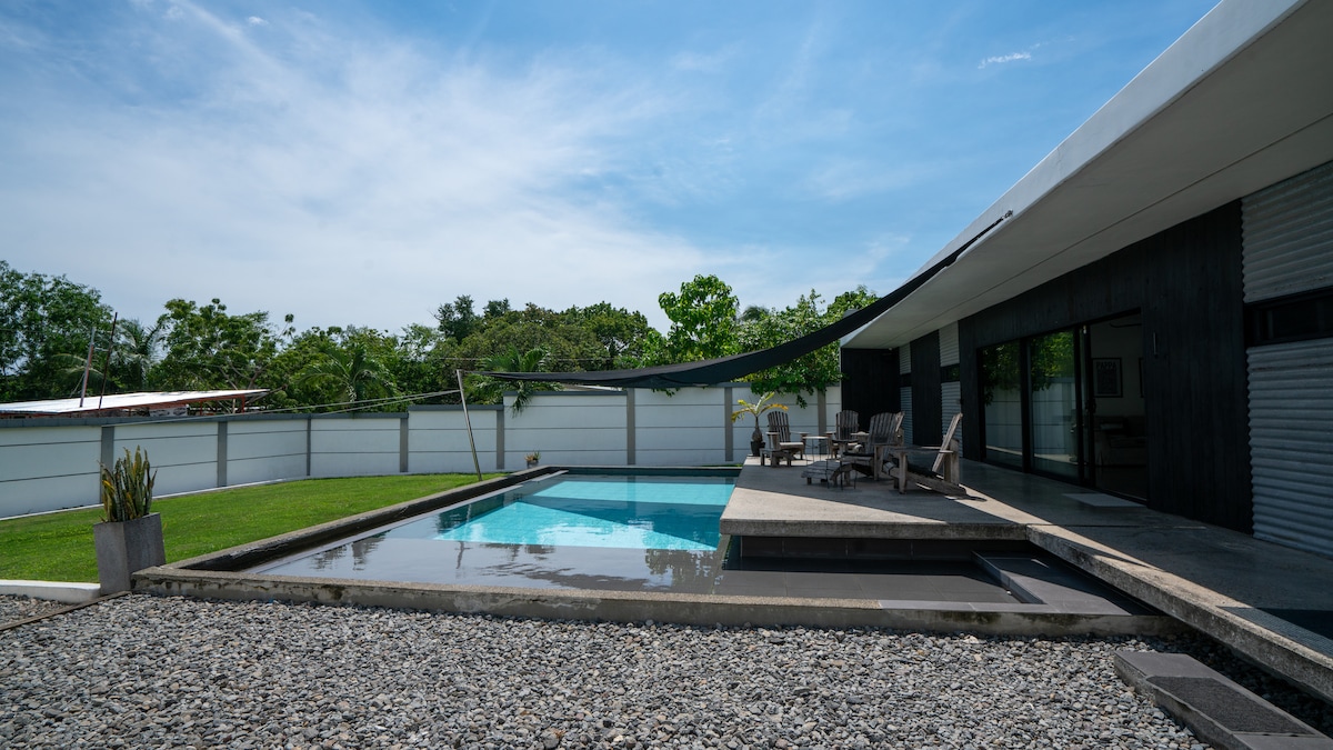 A private swimming pool is framed by a spacious deck and sleek outdoor furniture. Lush greenery is visible in the background, while the surrounding area is landscaped with gravel and grass. Natural light highlights the modern architectural features of the adjoining structure.