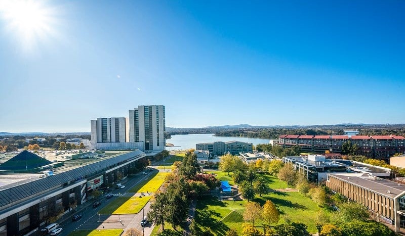 A panoramic view captures the landscape of Belconnen, featuring a serene lake bordered by greenery. The skyline includes modern buildings, with a hint of urban amenities visible. Sunlight brightens the scene, emphasizing the vibrant ambiance of the surrounding area.